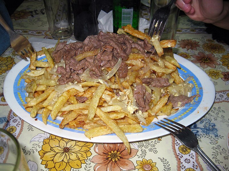 Chorrillana - a plate of fries with cut up meat on top, with three forks reaching out for it, set on a table with bottles in the background.