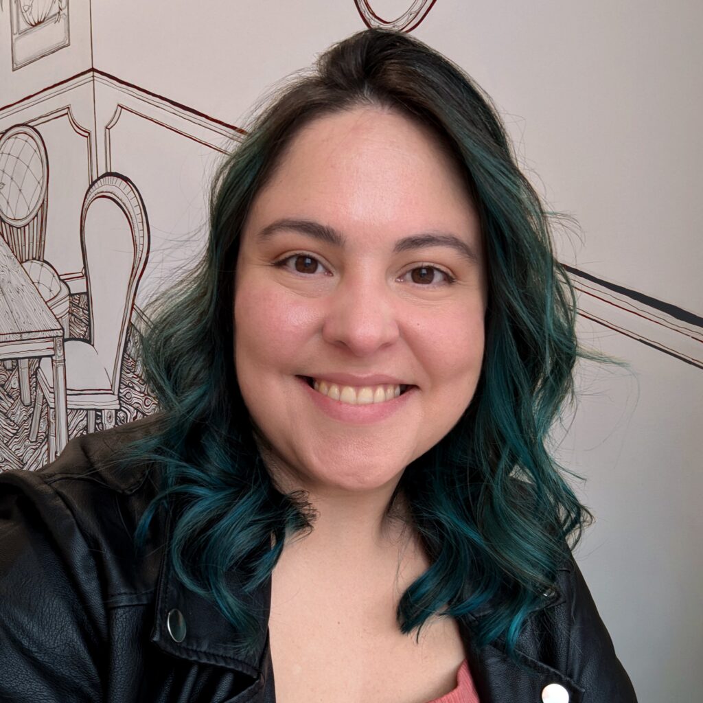 Leonor smiling at the camera. Her hair is curly down to her shoulders and dyed a dark teal. She wears a pleather jacket and rust-colored dress.