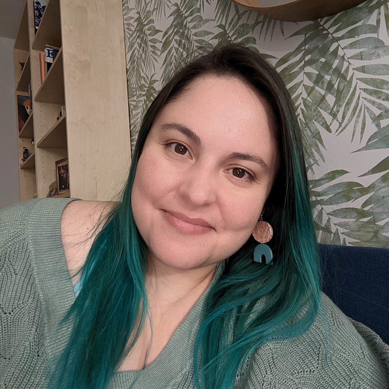 Leonor Soliz, Comfort Romance Author. A close-up photo of the author smiling at the camera, with vibrant teal hair.