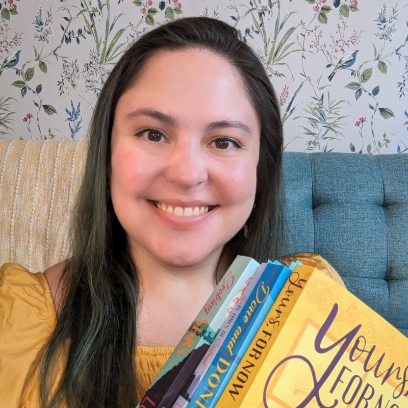 A photo of Leonor holding four of her books. She wears a yellow shirt, has long, straight brown hair with the ends dyed a blue-green color.